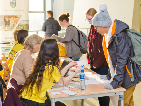 A new student leans over a desk where an access leader in a yellow tshirt is talking him through the UCD orientation programme